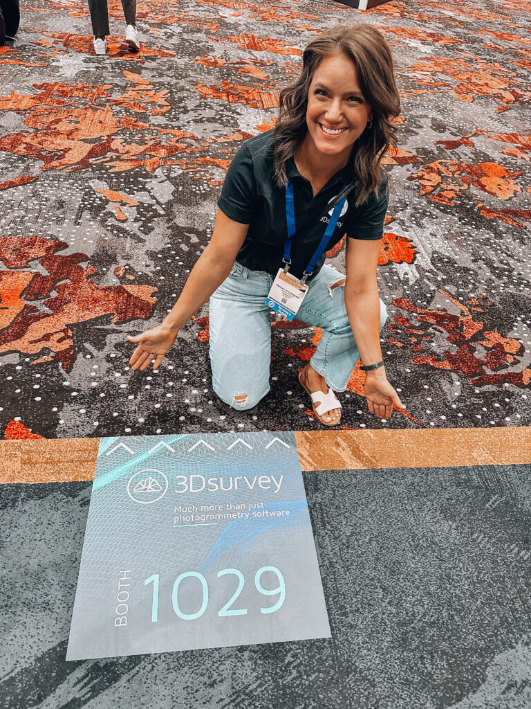 A smiling woman gesturing to a floor sticker that is promoting 3Dsurvey at the Commercial UAV expo